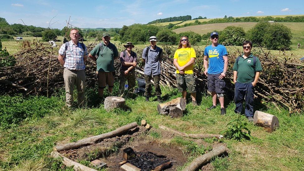 Seven men standing beside a fire pit on a sunny day surrounded by fields and trees