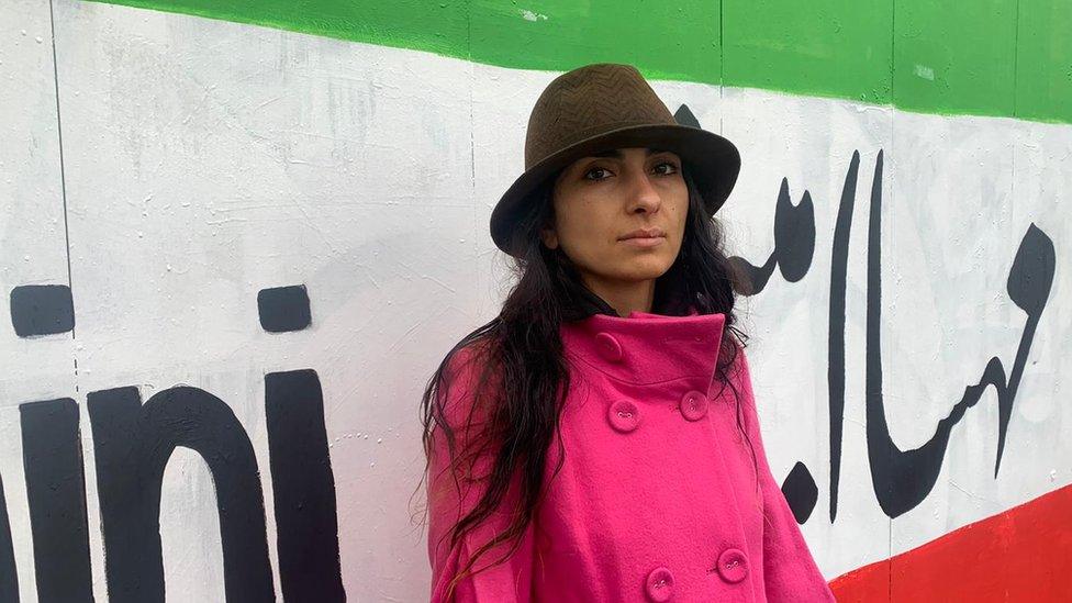 Artist Sahar Saki stands in front of her mural, and stares directly into the camera.