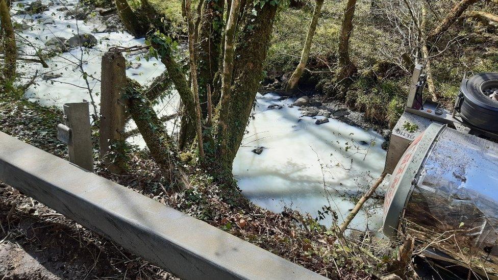 Dŵr Afon Dulais yn wyn a rhan tancer i'w weld yn gorwedd ar ei ochr ar y dde