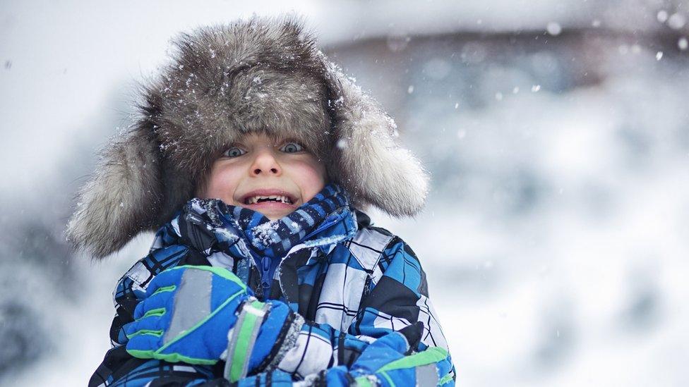 a child shivers wearing a furry hat and big warm gloves while outside in snowy weather