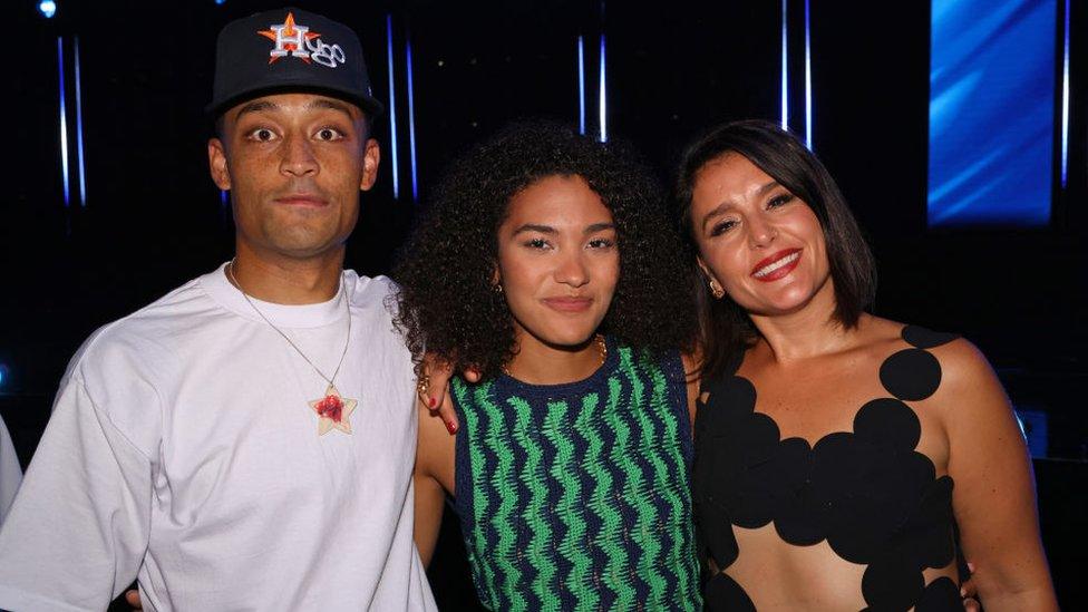 Loyle Carner, Olivia Dean and Jessie Ware attend The Mercury Prize 2023 awards show at Eventim Apollo on September 07, 2023 in London. Loyle wears a white T-shirt and black cap emblazoned with 'Hugo' and looks wide-eyed at the camera. Olivia, about a foot shorter, stands next to him, she wears her dark curl hair loose and smiles at the camera. She wears a green and blue zig-zag patterned dress. To her right is Jessie Ware, who tilts her head towards Olivia. She has short, straight dark hair and smiles at the camera, wearing deep red lipstick. Her dress is formed of connecting black spots. The trio are pictured inside at the ceremony and it is dark behind them with the exception of a blue screen