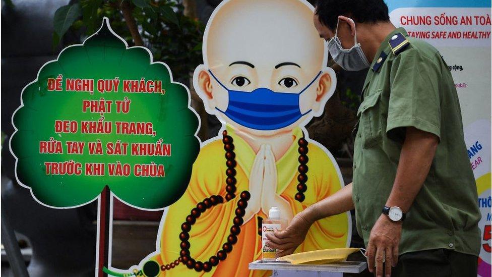 A local security officer disinfects his hands in Quan Su pagoda in front of a monk illustration wearing a face mask