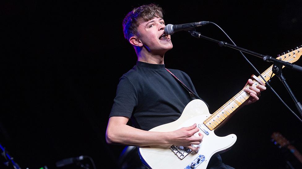 Musician Tom A Smith playing at a gig. He holds a white guitar and looks emotional, his mouth open wide as as he sings into a microphone. He's wearing a dark t-shirt and glancing out of the side of his eye at the camera.