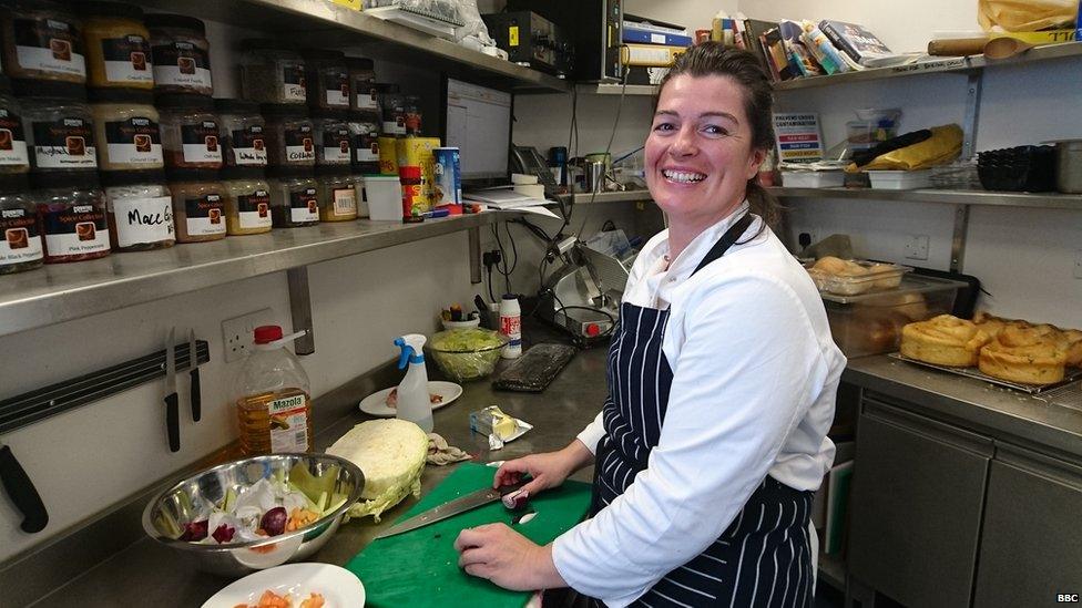 A lady smiles in the foreground, chopping food on a silver surface. Bread and spices fill the shelves around her