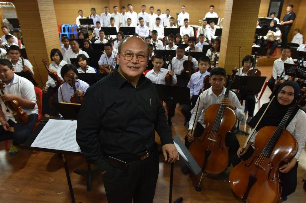 The conductor pictured in front of his orchestra in an indoor hall