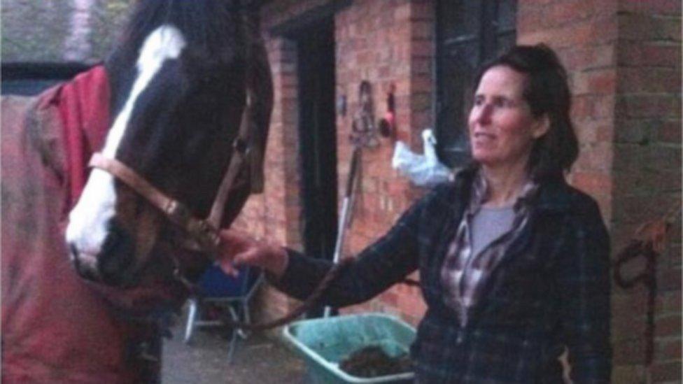 Woman with long dark hair holds a horse by its reins outside a stable