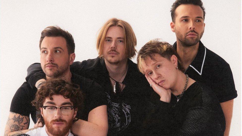 Five men pull nonchalant poses against a white background. In the middle is a man with chin-length dirty blonde hair, parted in the centre and wearing a black shirt open at the neck with a large silver pendant. He's got his arm around a man with short dark hair, who's looking off to the side, with short dark hair. On the centre man's left a man with short, dirty blonde messy hair has his cheek resting on one hand. He wears a thick silver chain and looks bored. Behind him a man in black shirt, with short dark hair and dark stubble, looks slightly off to the side. A final man, the only one wearing white, with glasses, curly dark hair and dark beard, sits in front of them.