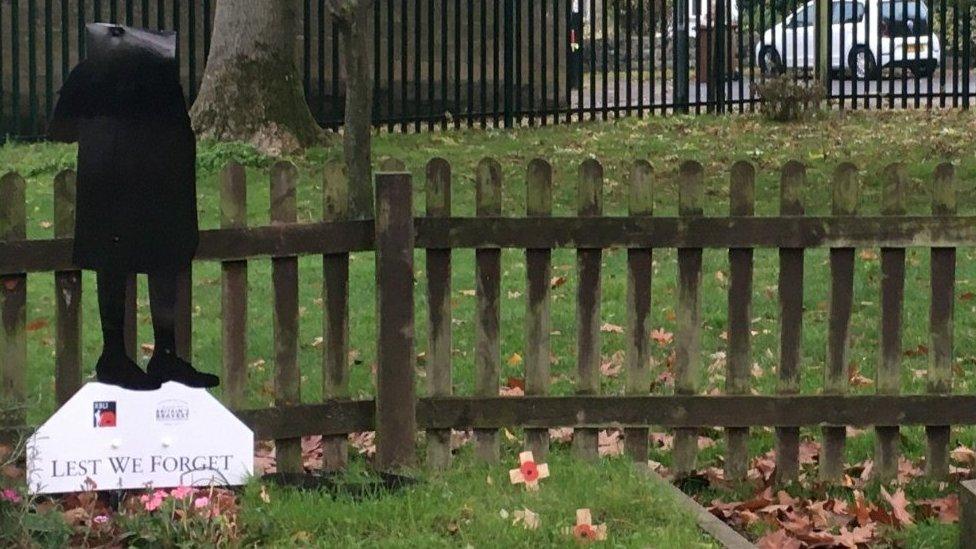 Female metal soldier figure with no head standing on a sign that reads "Lest We Forget"