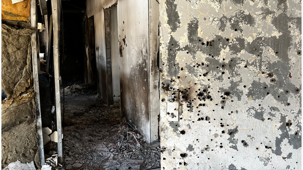 A metal door is punctured with countless bullet holes around the lock. It leads to a scorched corridor filled with debris