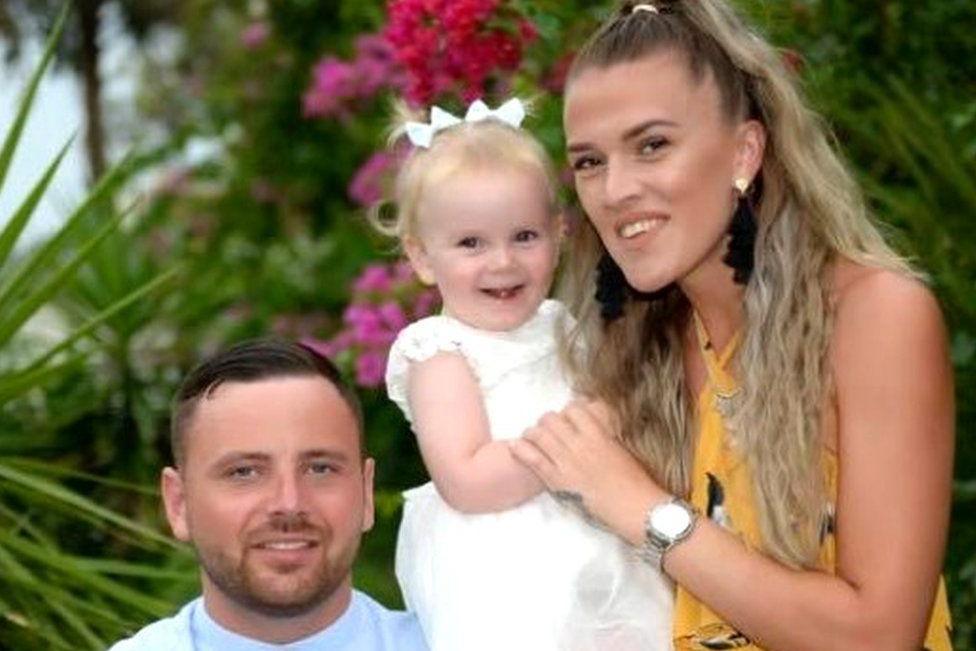 Family picture of a man seated wearing a blue shirt, a smiling woman standing next to him holding a smiling toddler in a white dress with matching bows in her hair, standing in front of flowers and palm trees