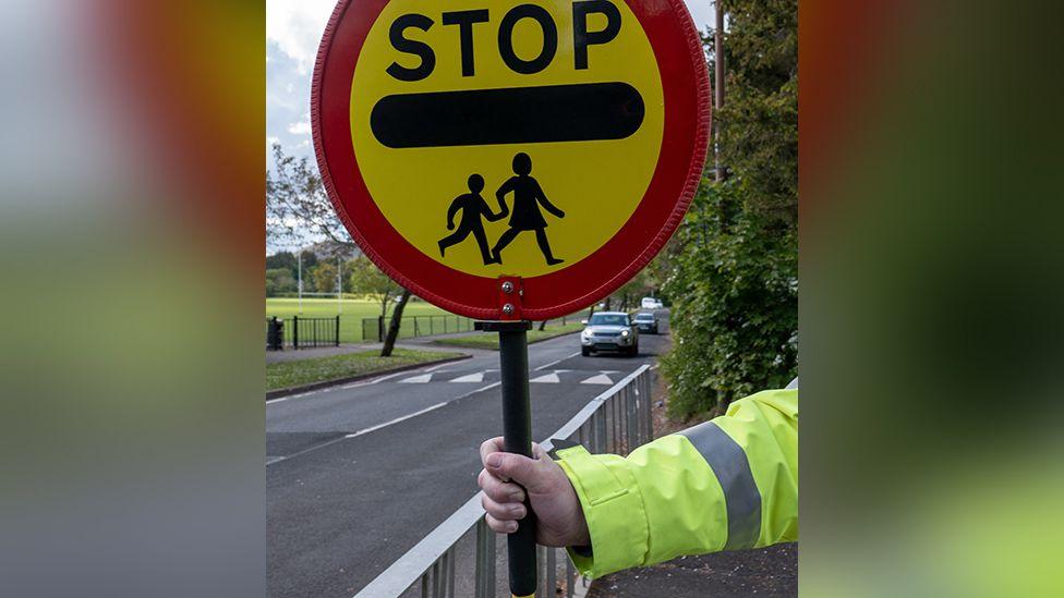 The arm of an anonymous lollipop warden, wearing a yellow hi-vis coat at a school crossing. The warden is holding a sign telling drivers to stop for children.