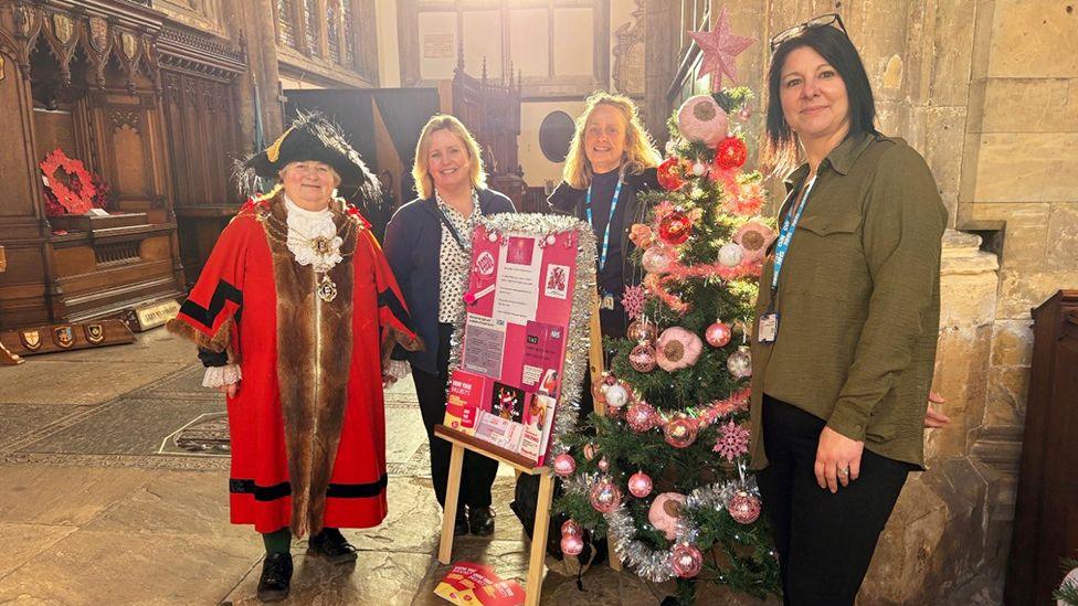 A number of NHS staff stand in front of the tree with the Lord Mayor in her red ceremonial outfit with chain and black tricorn hat. The tree has the knitted "boob-les" and has pink tinsel and a star on top