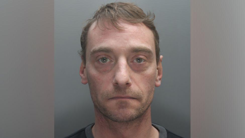 A police mugshot of Paul Wilson, who has short light brown hair, light stubble and blue eyes, and stares into the camera with a blank expression