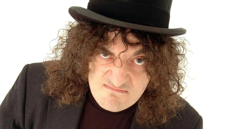 A man in a black blazer and top hat with long, brown curly hair - he raises one eyebrow and wears a grumpy expression