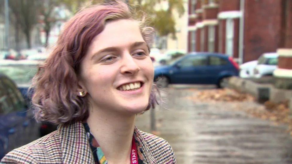 Alexa Moore smiling in a checked coat, pictured on a street with homes and a blue car in the background