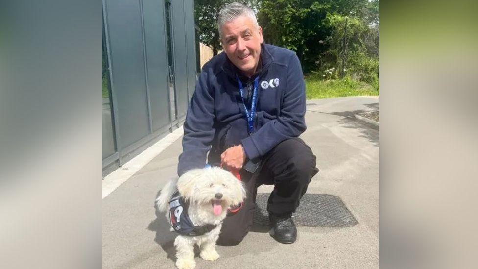 A small fluffy white dog next to a man in a blue jacketkneeling beside it