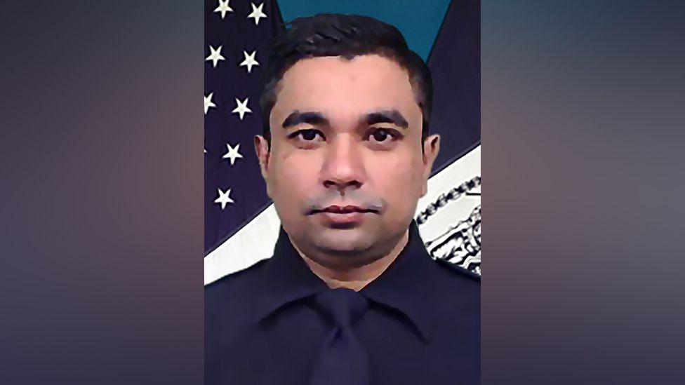 Didarul Islam, shown from the shoulders up, in an official police portrait. He is in a dark blue shirt and tie. In the background is part of an American flag.