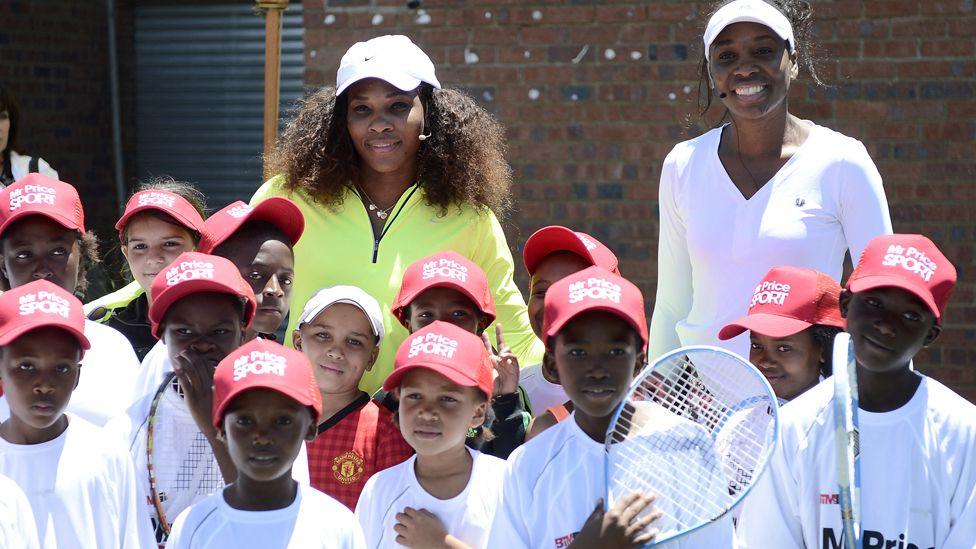 US tennis player Serena Williams (left) in a yellow top and white cap flanked by her sister Venus in a white top and cap pose with children in red caps and white T-shirts, some holding rackets after a two-hour tennis clinic at the Arthur Ashe Soweto Tennis Centre - November 2012.