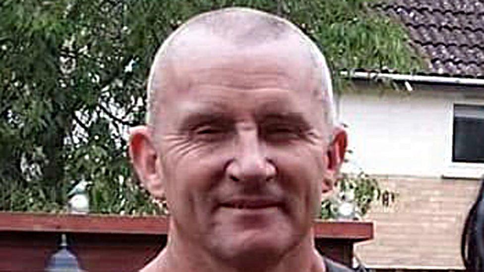 Bryan Field with a shaved head is standing in his garden with a shed behind him.