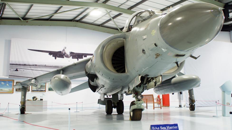 A white aircraft in a museum with a BAe Sea Harrier sign below it