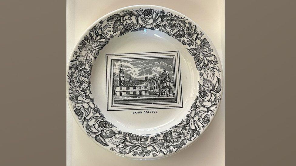 A white plate is decorated with a view of Caius Court in its centre, in black, and the words "Caius College" printed beneath. Around the edges are black drawings of flowers and foliage