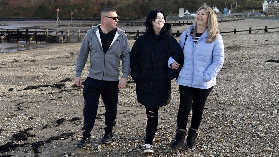 Brian Buckle, along with his daughter Georgia and his wife Elaine, walk along a beach on a sunny-ish day. Brian is wearing a tracksuit top and dark glasses, Georgia is wearing black and Elaine has a lilac puffer jacket