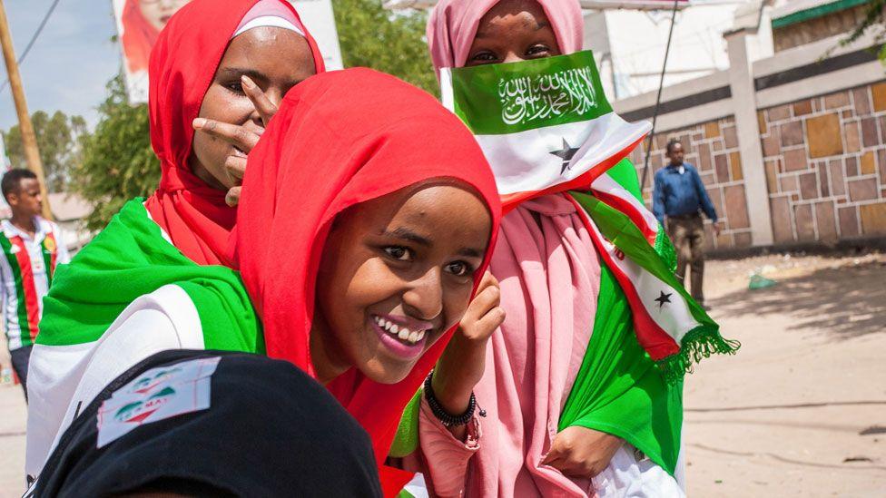 Three young women smiling wearing red and pink headscarves and holding a Somaliland flag and other regalia - Hargeisa, 2018