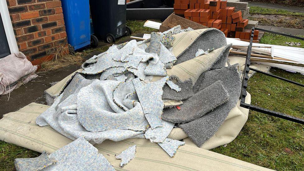 A pile of ripped up grey carpet in a garden outside a house. Sandbags can be seen against the step and door of the house. A pile of bricks and two wheelie bins are in the background.