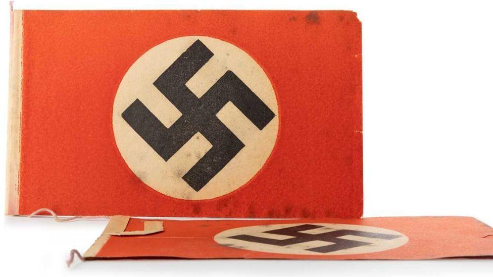 Nazi bunting photographed against a white background. Two individual Nazi flags are on display, one lying flat in front of another upright facing the camera. The flags are red with a white circle in the middle containing a black swastika. 