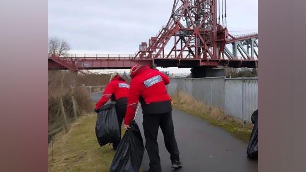 A man and a woman picking up litter and putting it into black bags while walking along a riverside path, with Middlesbrough's Newport Bridge in front of them.