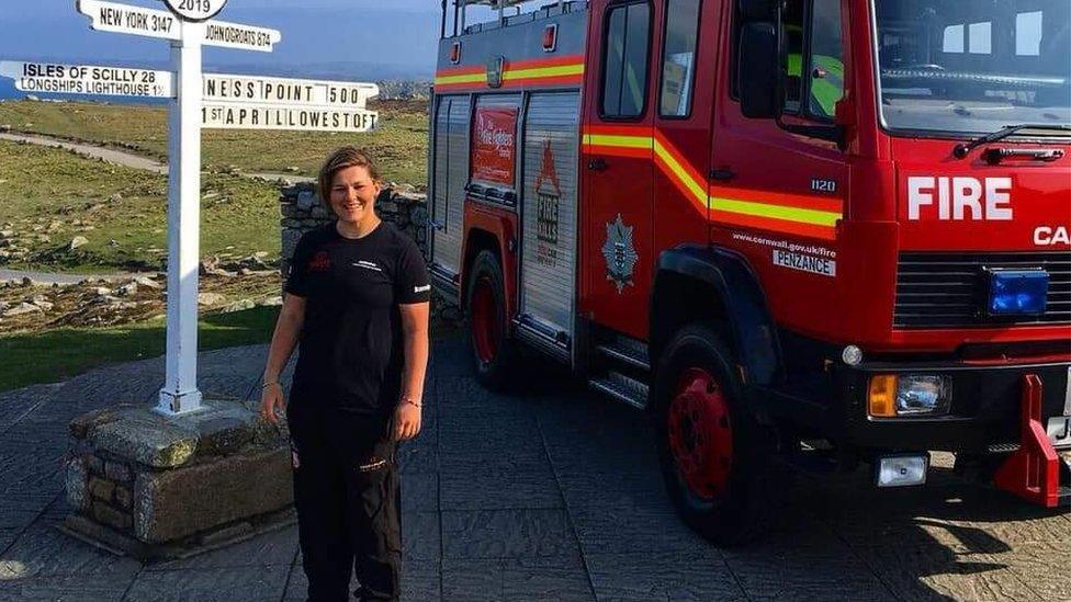Sasha Acheson wearing all black and standing beside the landmark sign at Lands End, posing for a picture. Behind her there is a large red fire engine with hi-vis decals.
