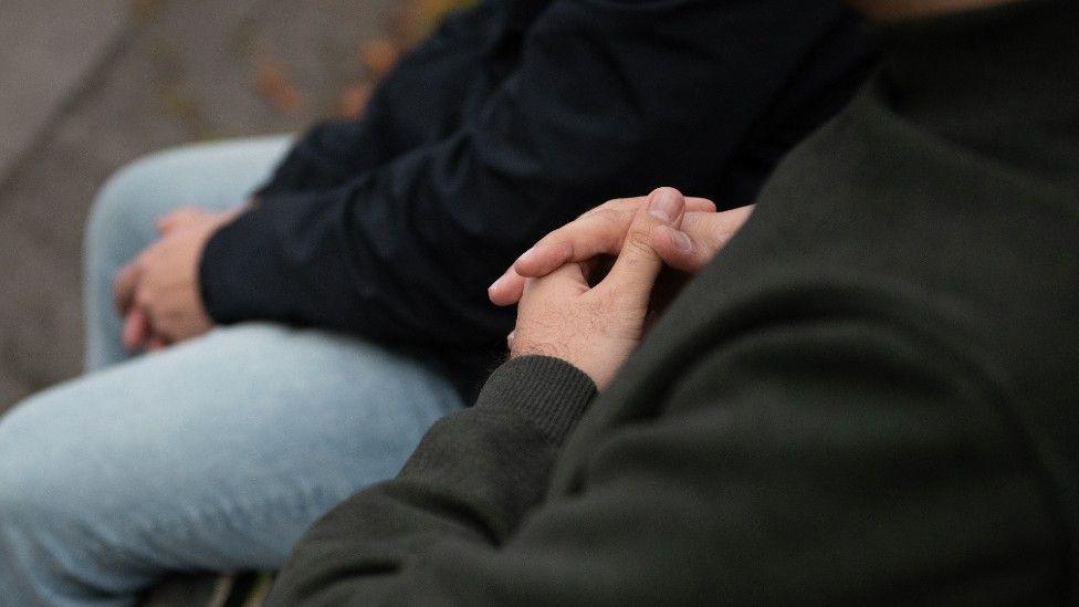 A close-up of two men sitting on what appears to be a park bench. Their faces are not visible but we see the hands of one of the men clasped together.