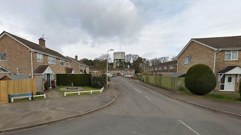 Road leading into close with houses each side and along the end - a large tower stands in the centre behind the houses.