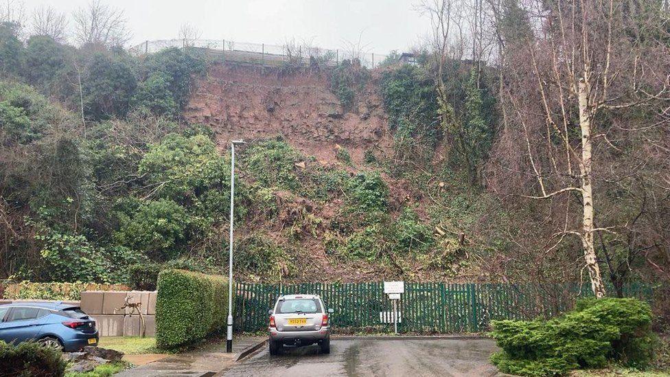 View down a short road to the cliff face, showing a mass of vegetation and mud at the bottom of a bare rocky cliff