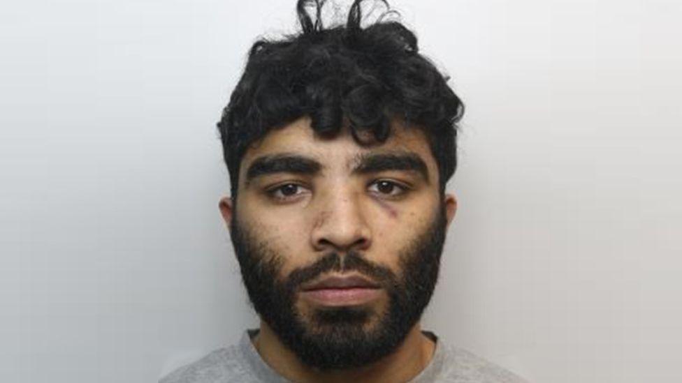 A police mugshot of Hassan Jhangur. He has dark facial hair and dark, slightly curly hair.