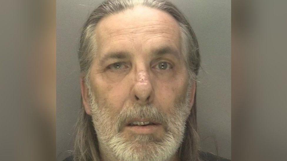 A mugshot of a man in his early 60s. He looks at the camera and is in front of a grey background. He has grey shoulder-length hair and a white beard.