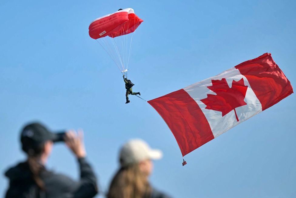 A large Canadian national flag hangs from a parachutist as he lands ahead of the Canadian commemorative ceremony marking the 80th anniversary of the World War II "D-Day" Allied landings in Normandy, at the Juno Beach Centre near the village of Courseulles-sur-Mer, in northwestern France, on June 6, 2024.