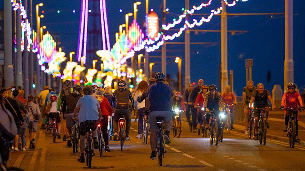 Blackpool prom at dusk with dozens of cyclists seen at a distance riding under Blackpool Illuminations