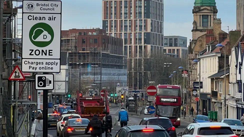 A congested road pictured in the daytime, with a clean air zone sign on the left