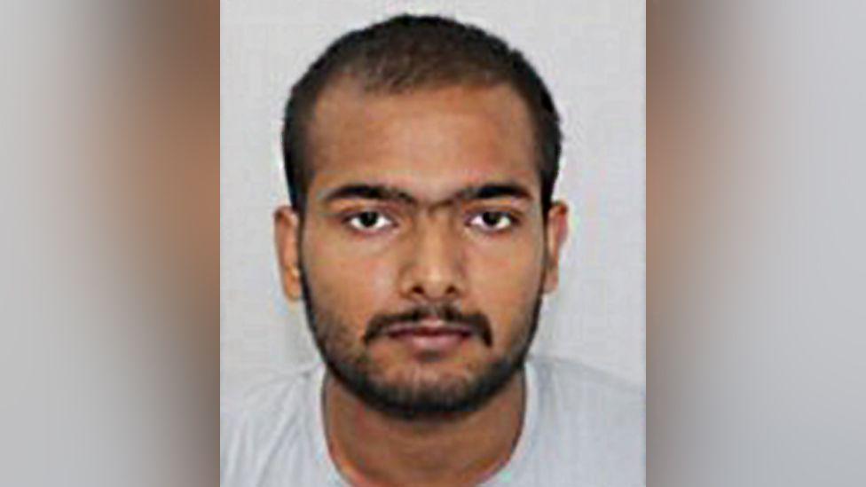 Custody photo of Pankaj Lamba against a grey background. He has dark facial hair, brown eyes and is looking at the camera.