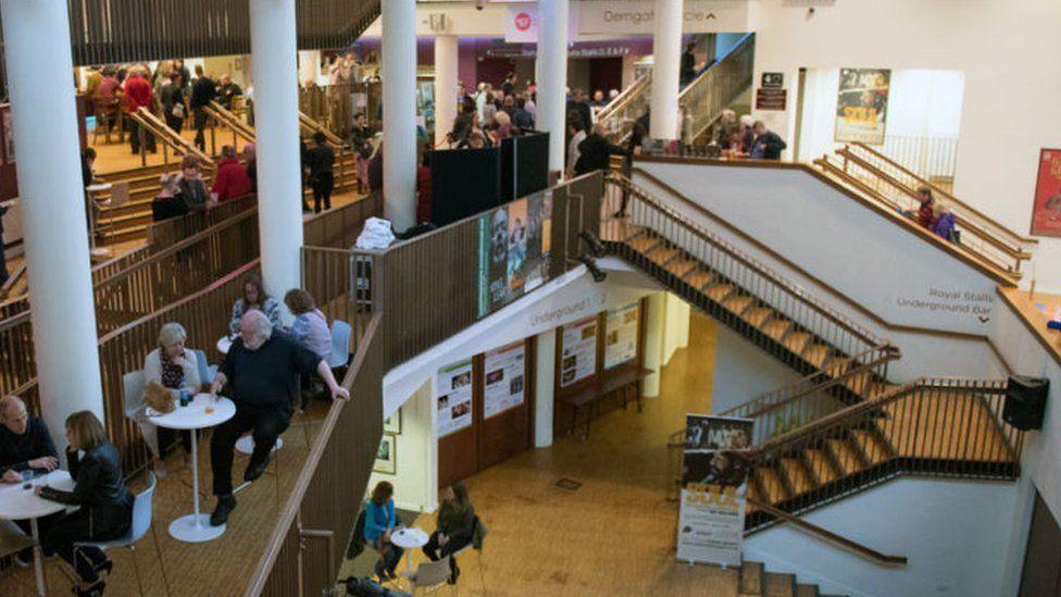 Foyer of the Royal and Derngate, showing large white pillars on the upper floor and gold-coloured stairs down to the ground floor and underground levels. People are sitting at small tables to the left of the picture enjoying pre-performance drinks.