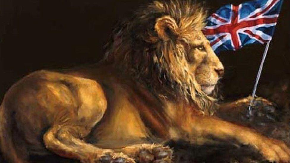 A lion lying down on its side, the underneath of both its rear paws can be seen with its front left holding a pole with a Union Jack flag.