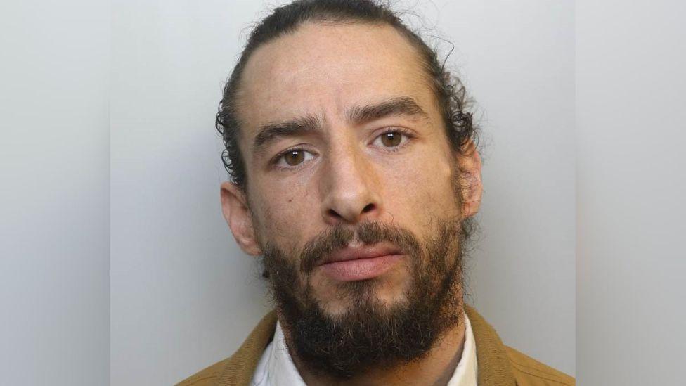 A custody picture of Cody. He has long dark hair pulled back from his face, dark eyebrows and a dark beard. He wears a white collared shirt and what appears to be a tan-coloured jacket.