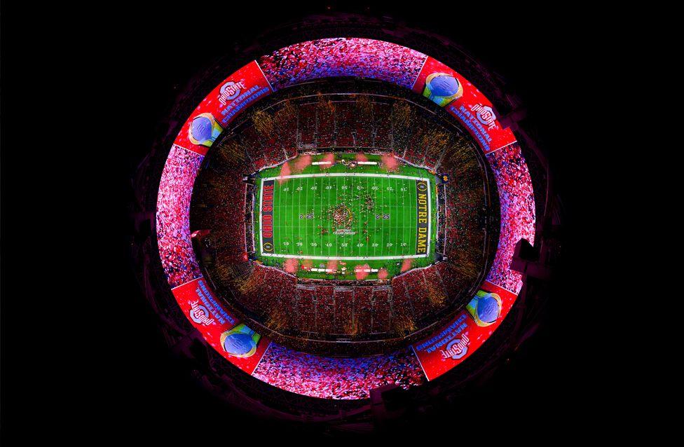 A fisheye view of a brightly lit football stadium packed with fans. The green field is at the centre, surrounded by vibrant red and blue banners displaying team names and championship graphics.
