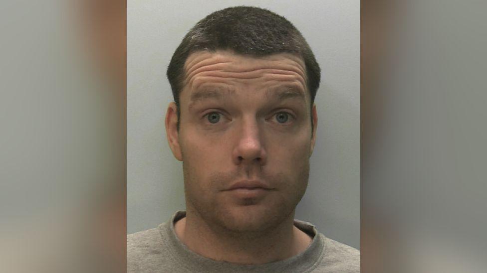 A mugshot of Daniel Lynch. He is raising his eyebrows and his eyes are wide open. He has short dark brown hair and is unshaven. He is wearing a pale brown sweatshirt.
