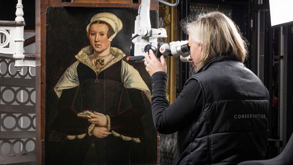 A head and torso portrait of a Tudor woman believed to be Lady Jane Grey.is wearing a white linen cap, a white lacework open collar, a gold necklace looped around her neck. Her dress has black and has a white scarf over its shoulders. Her hands are in front of her waist and the left had is holding a book. Rachel Turnbul is on the right examining the portrait with conservation equipment and her black gilet says conservation. 