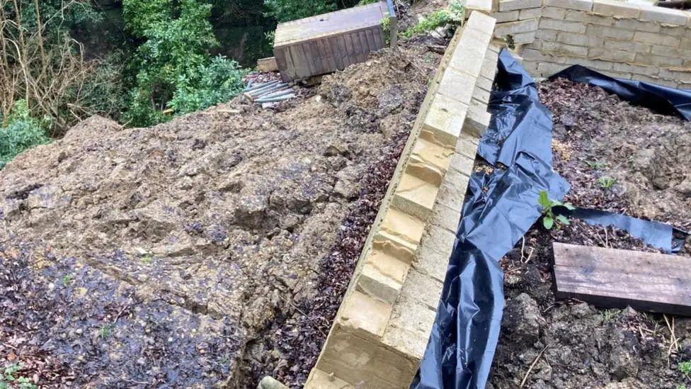 Part of a low, pale brick wall that has collapsed due to the landslip with mud seen on a downward slope behind the wall and plastic sheeting on the other side.