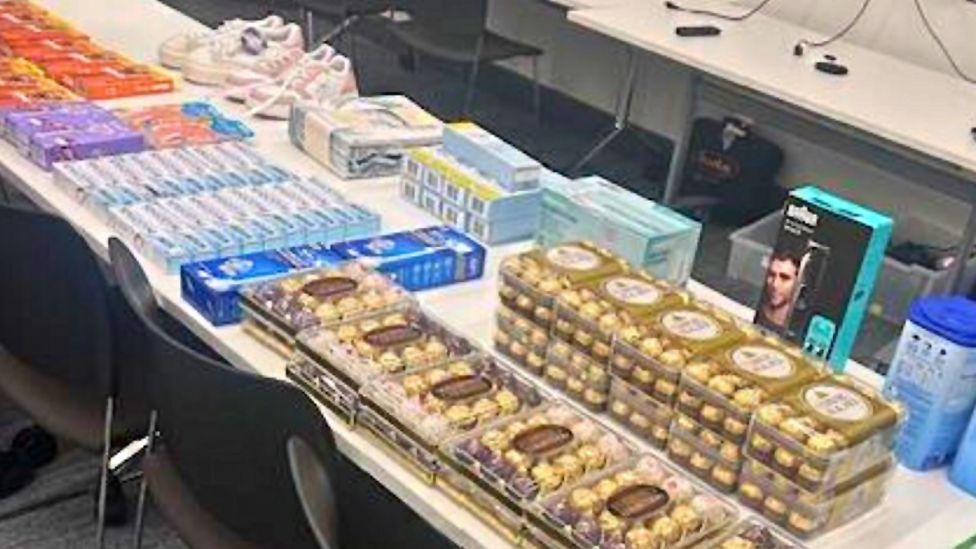 A table of items including confectionary, trainers, toothpaste boxes and other items all laid out on a white table.