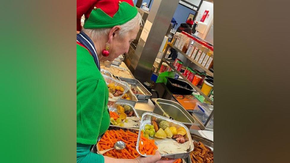 A woman with short grey hair is wearing a red and green elf costume. She is holding a tray filled with a Christmas dinner. There are plates around her on a table that contain vegetables and meat.