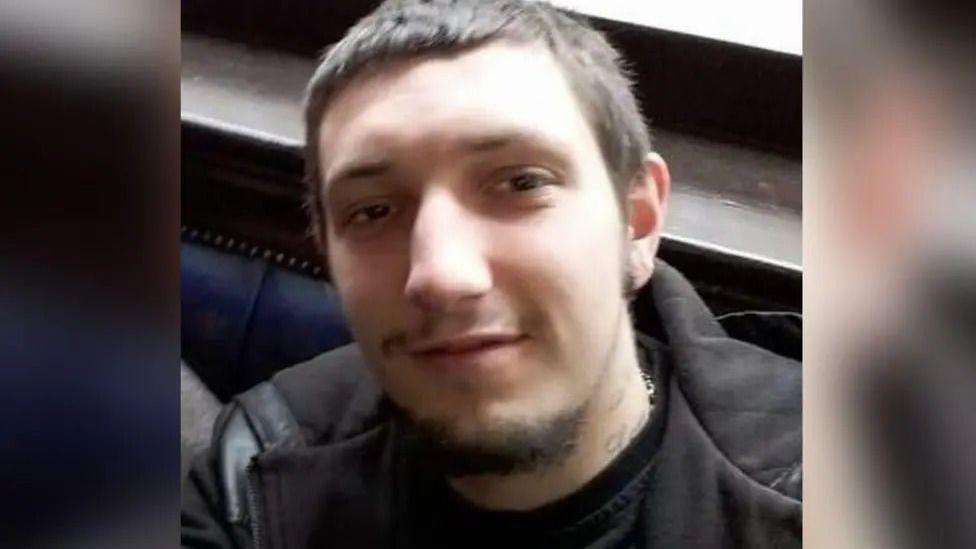 A young man in his twenties, wearing a black jacket. He has short brown hair and a short beard.